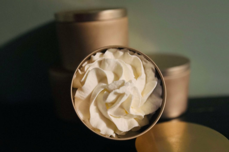 Close-up of a decorative cream or lotion in a round, gold-topped tin, resembling a flower. In the background, additional gold tins are partially visible, creating an elegant display for skincare or beauty products.