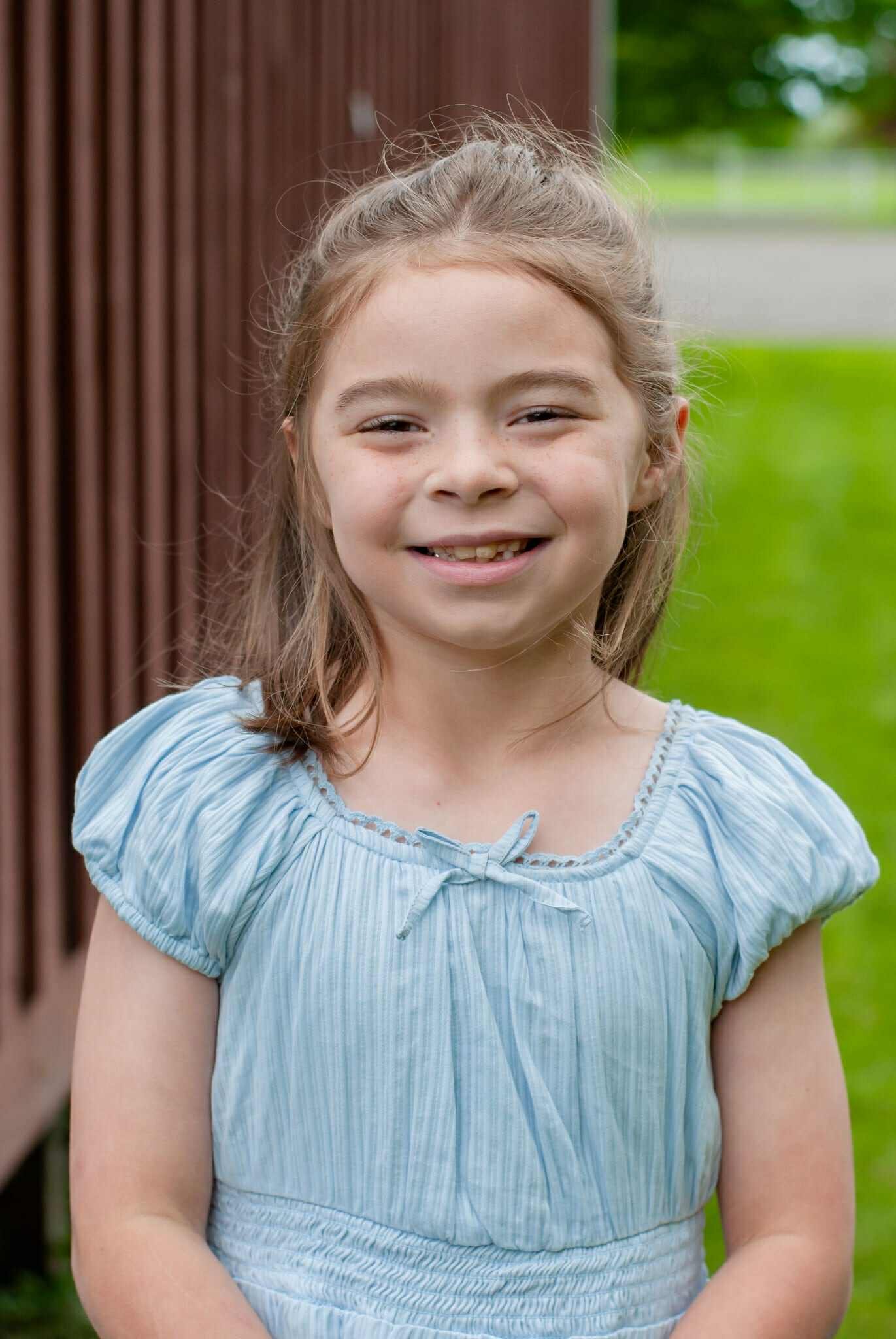 Smiling young girl wearing a light blue dress, standing outdoors with green grass in the background and a wooden structure behind her.