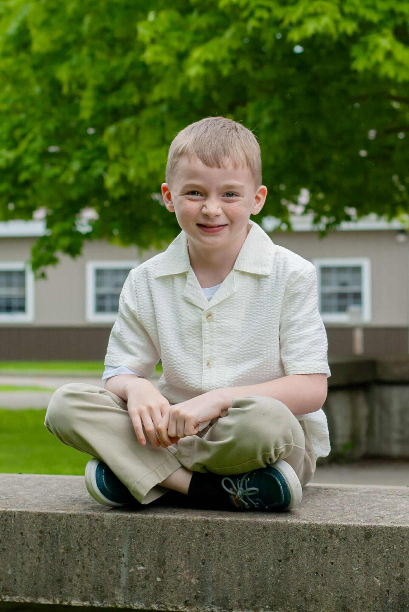Smiling young boy sitting cross-legged on a stone ledge, wearing a light-colored short-sleeve shirt and beige pants, with a green leafy background.