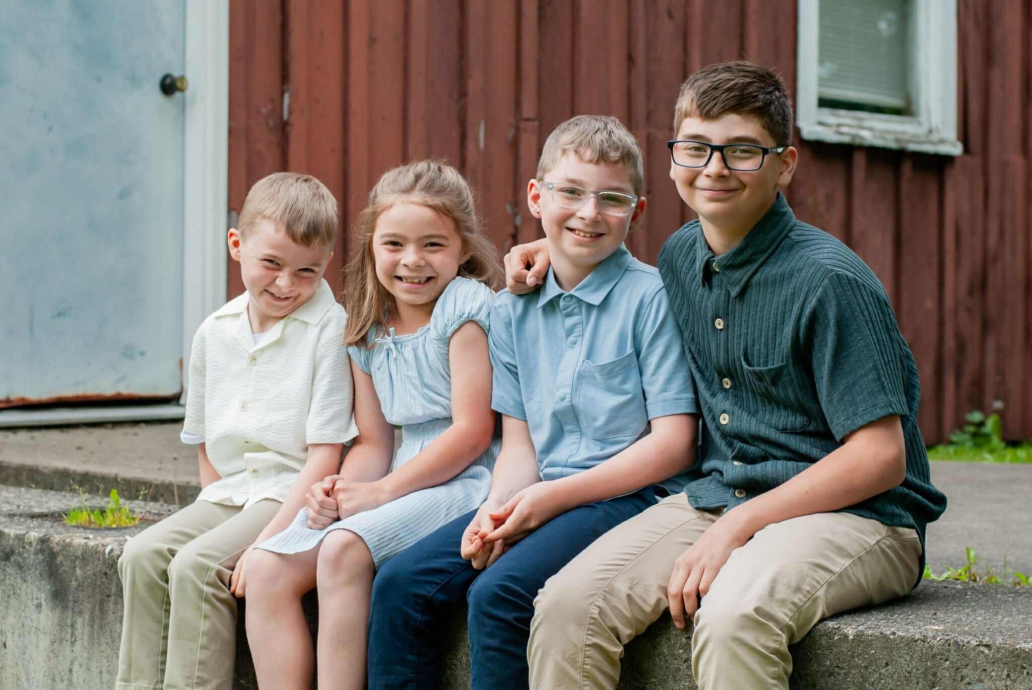 Four children sitting on a concrete step outside a building, smiling and dressed in casual summer clothing. The background features a rustic wooden wall and a door, creating a warm, inviting atmosphere.