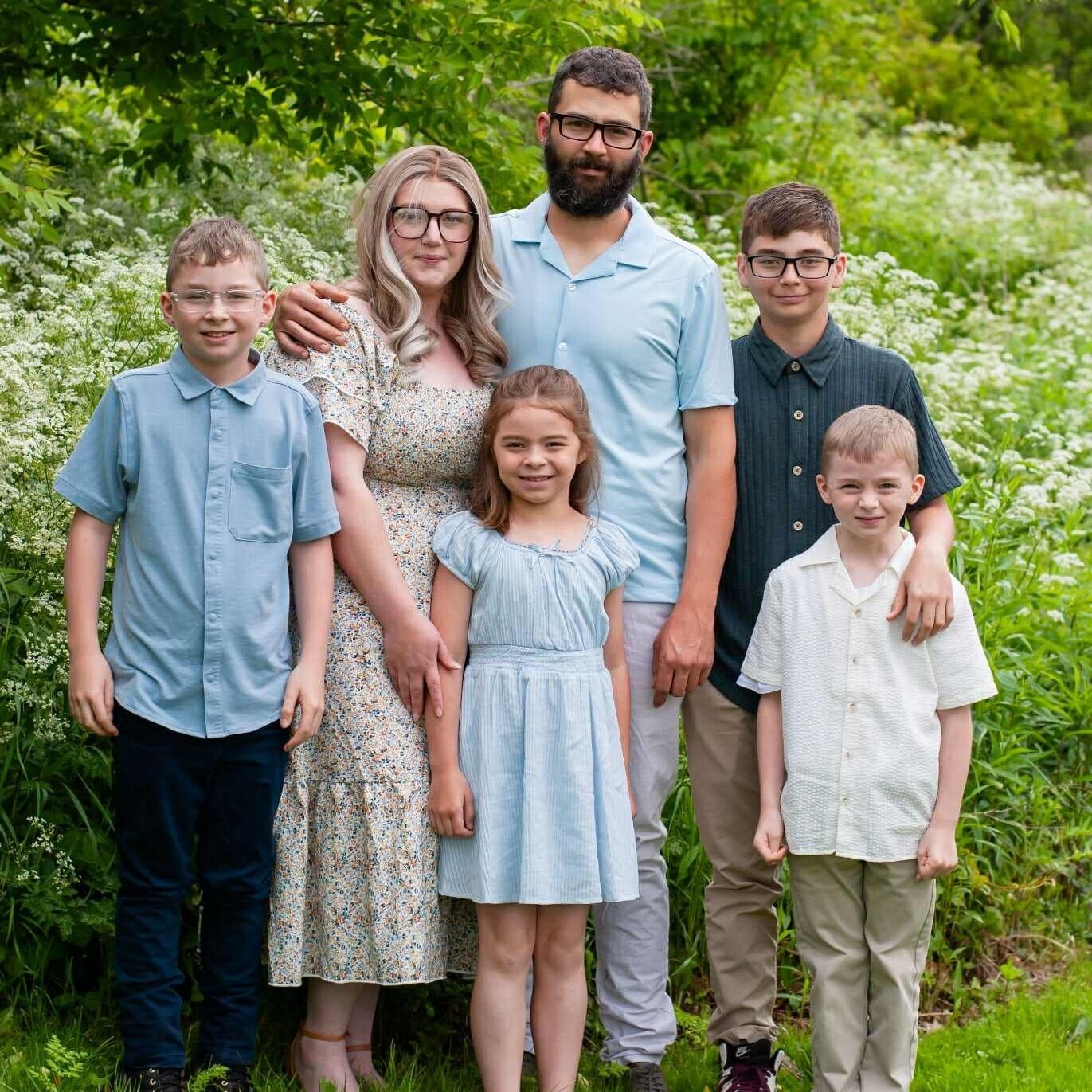A family portrait featuring five members standing together in a lush green garden. The parents, a mother in a floral dress and a father with a beard, are flanked by four children in light-colored clothing, all smiling and posing for the camera.