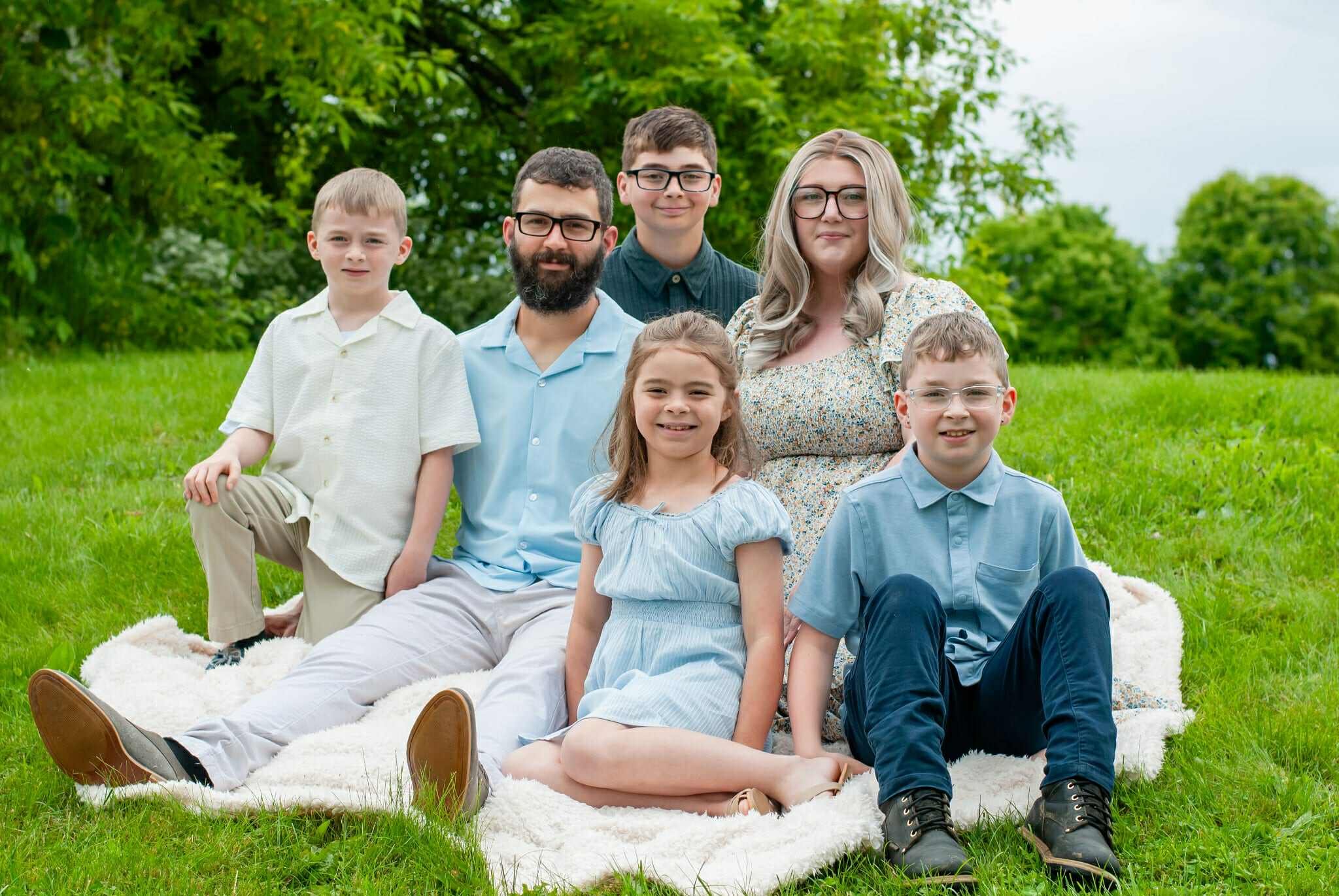 Family portrait featuring six members sitting on a blanket in a green park. The group consists of three children and three adults, with a backdrop of lush trees and a cloudy sky. The children are dressed in light-colored clothing, while the adults wear casual attire, creating a warm and inviting family atmosphere.