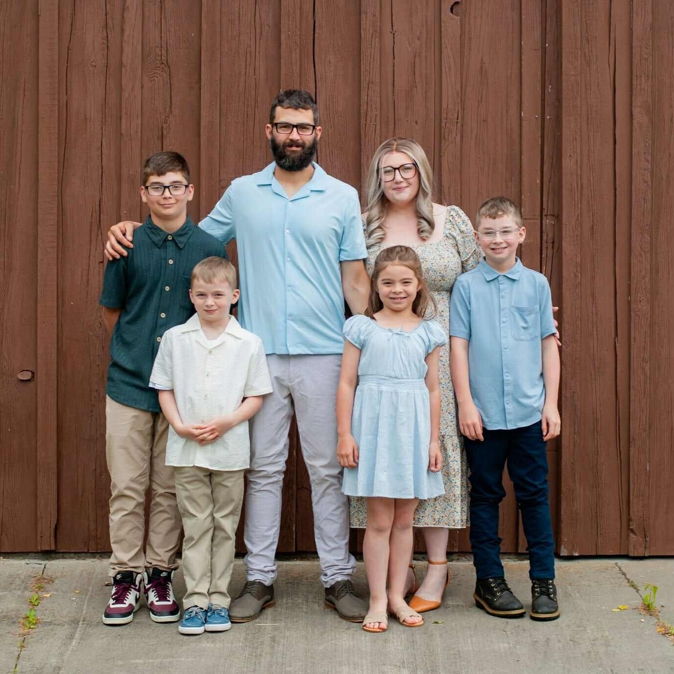 A family of six poses for a photo in front of a wooden backdrop. The group includes four children and two adults, all dressed in casual, coordinated outfits with shades of blue and neutral tones. The setting conveys a warm and friendly atmosphere, perfect for family gatherings or portraits.