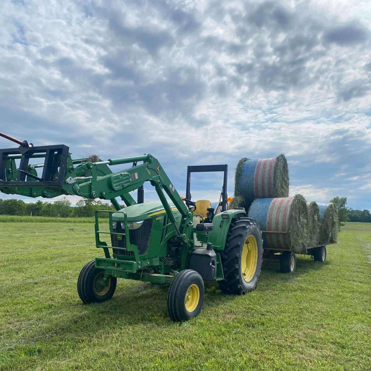 A green tractor equipped with a loader is lifting hay bales on a sunny day in a grassy field, with additional bales loaded on a trailer behind it.