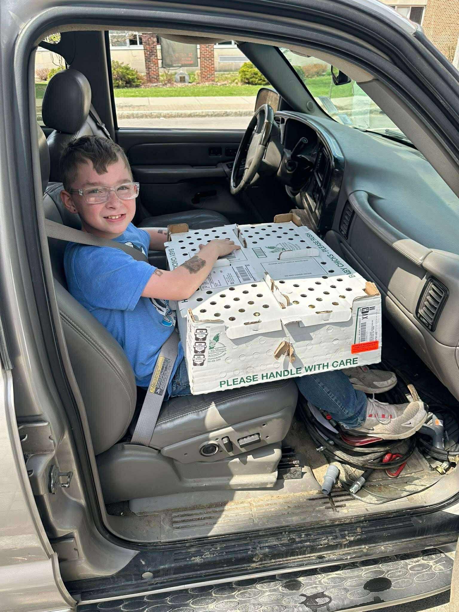 Young boy sitting in the driver's seat of a truck, smiling while holding a box labeled "PLEASE HANDLE WITH CARE." The interior shows a slightly messy floor with various items, capturing a casual moment during a sunny day.