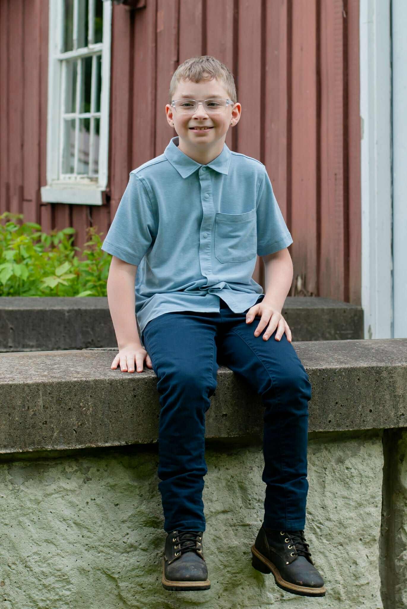 Smiling boy wearing glasses and a light blue polo shirt sitting on a stone wall, with a rustic red barn and greenery in the background.