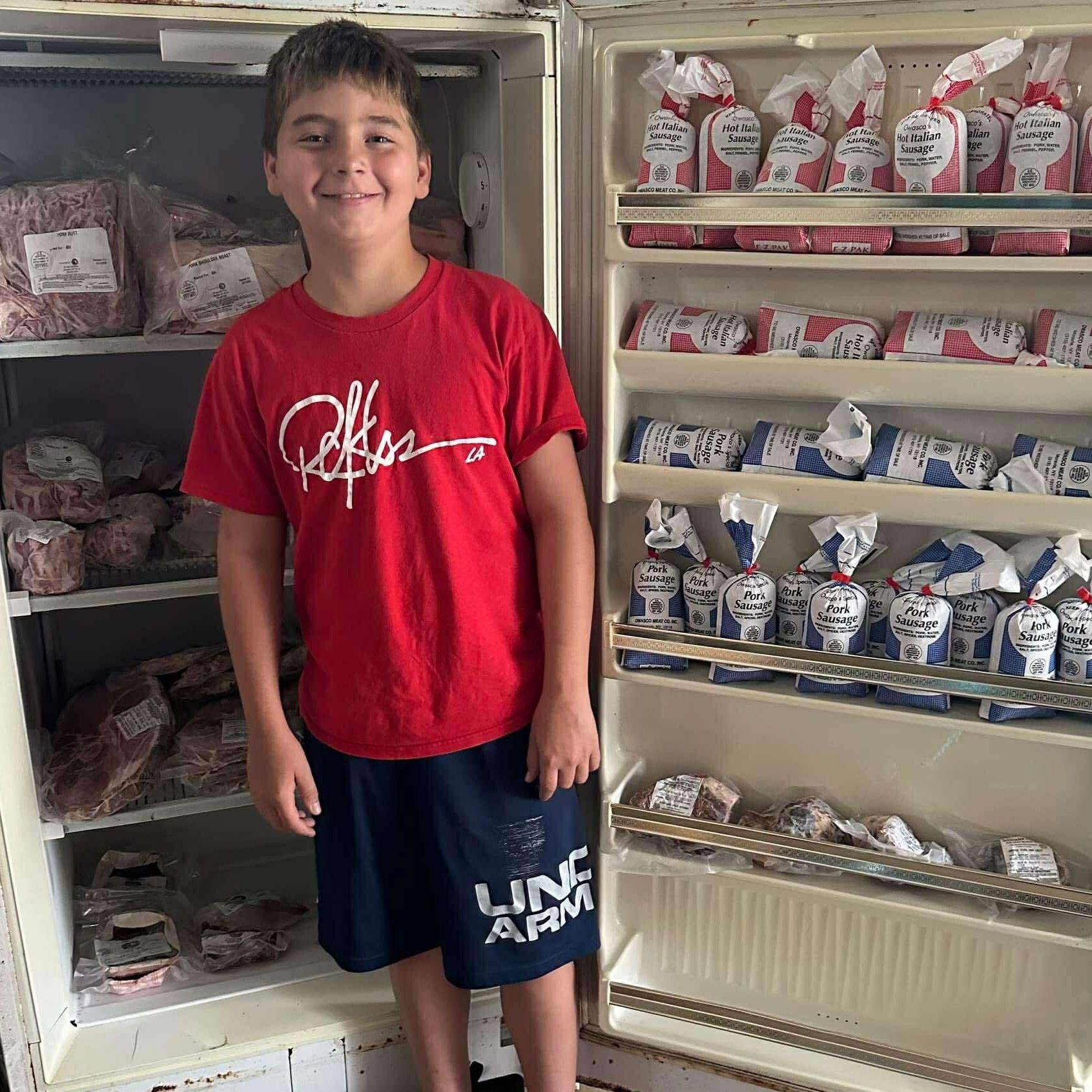 Smiling boy in a red shirt stands next to an open freezer filled with packaged meats, including various types of sausages and cuts of pork. The freezer showcases a variety of labeled products, highlighting a selection of quality meat options.