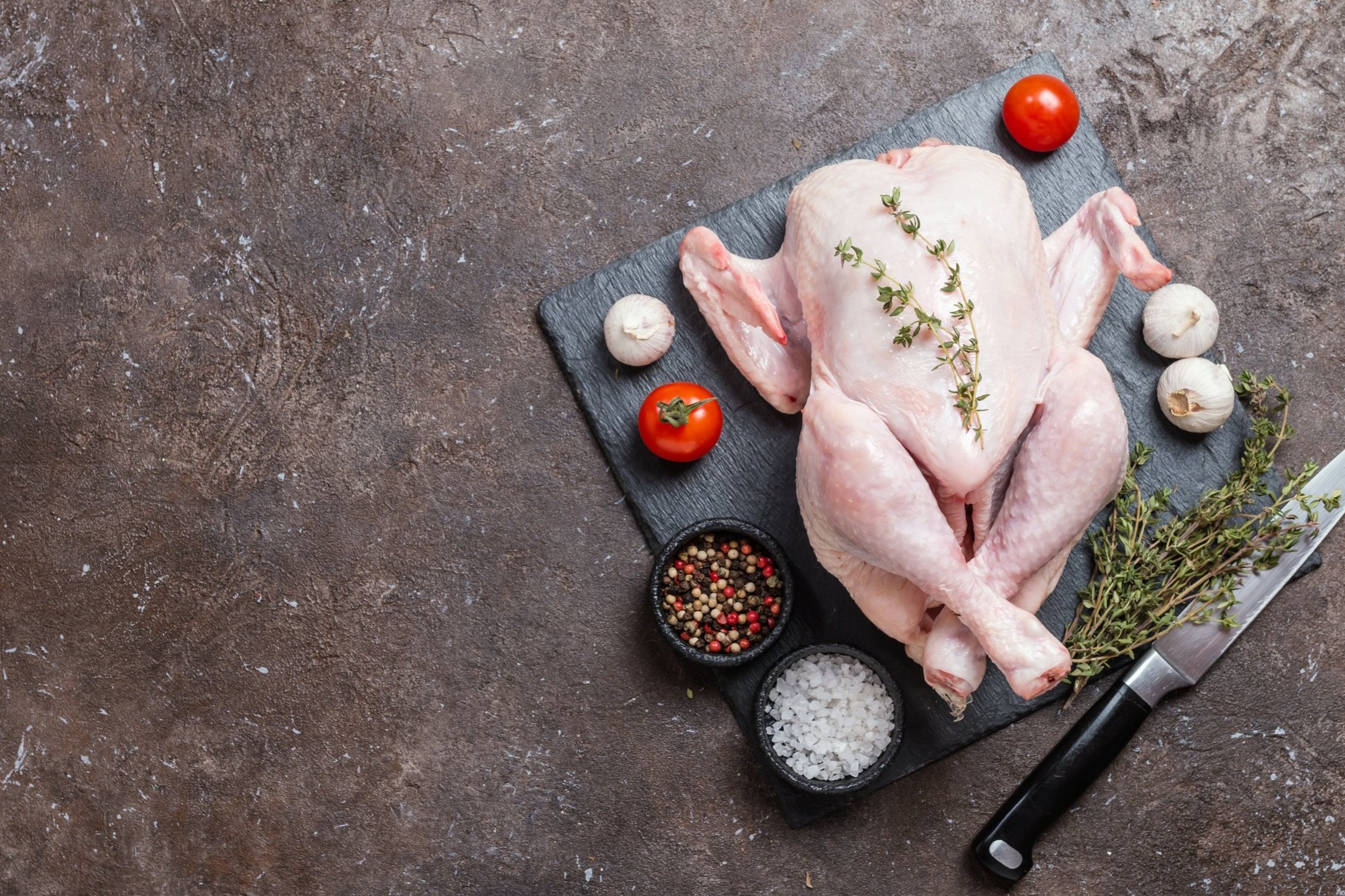 Whole raw chicken on a black slate cutting board, garnished with fresh thyme, surrounded by cherry tomatoes, garlic cloves, and bowls of mixed peppercorns and sea salt, with a sharp kitchen knife nearby.