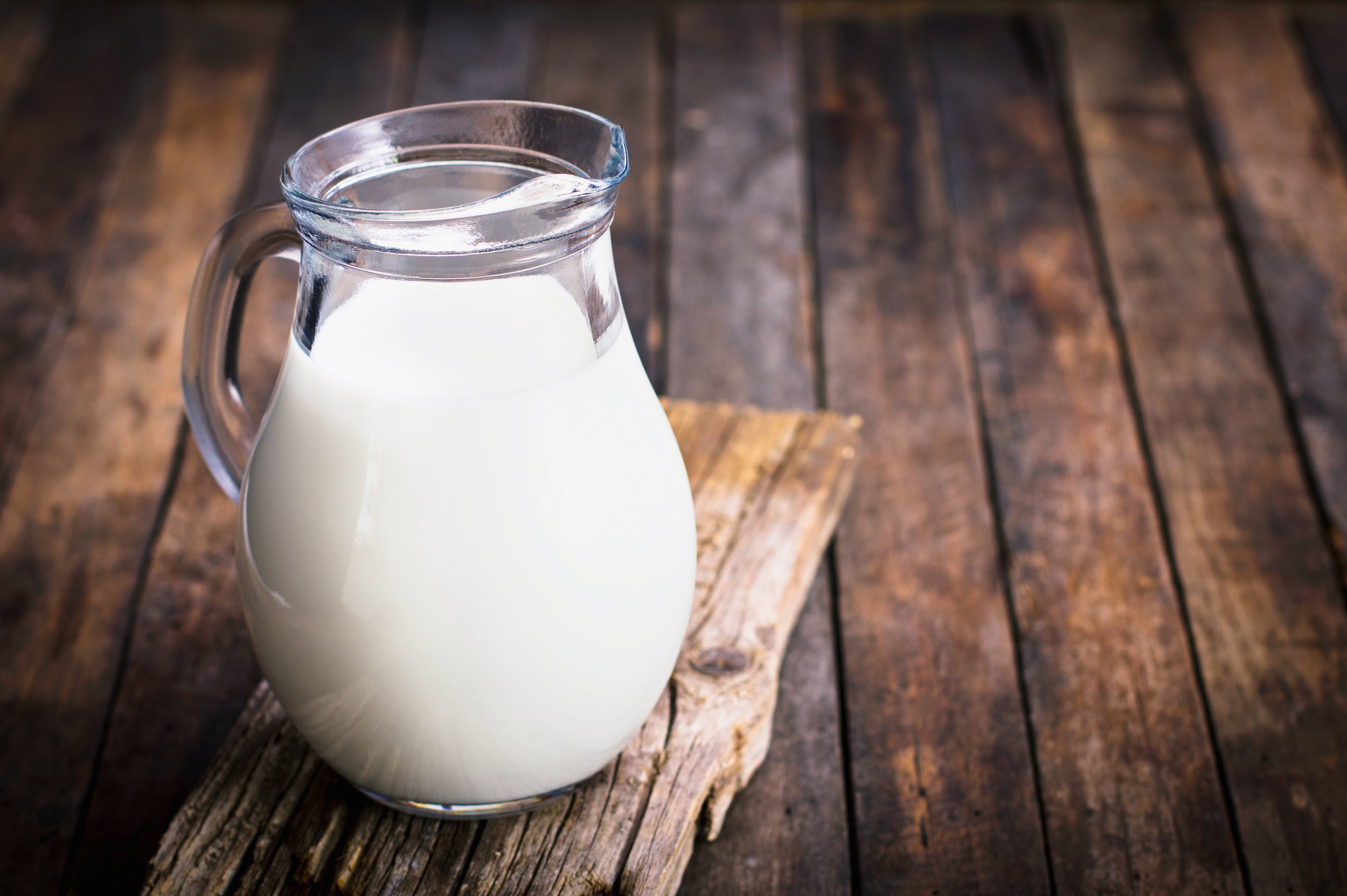 Glass pitcher filled with fresh milk placed on a rustic wooden surface, showcasing a simple and natural dairy product.