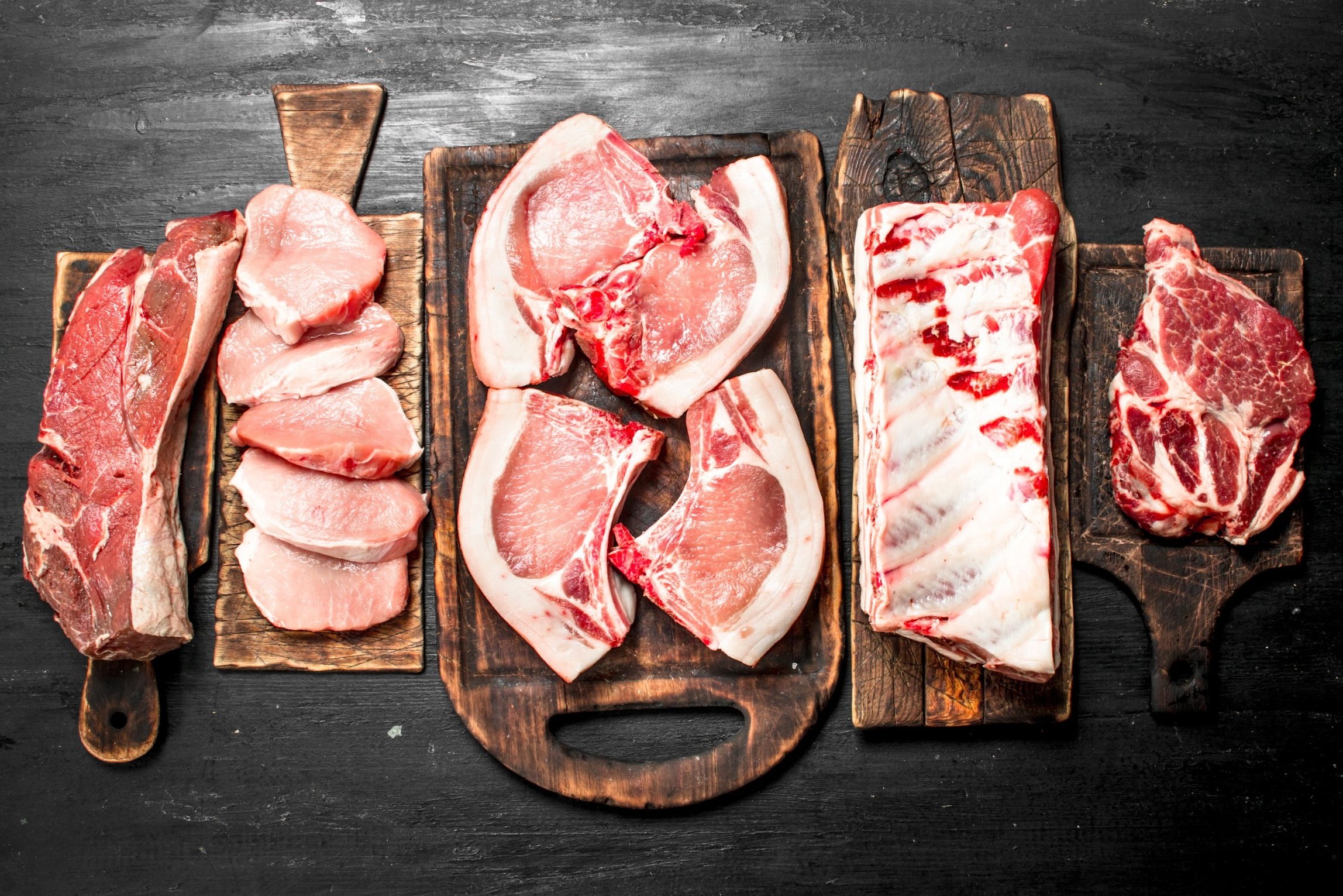 Assorted cuts of fresh pork meat displayed on wooden cutting boards, including pork chops, tenderloin, and ribs, arranged on a dark background for a culinary presentation.