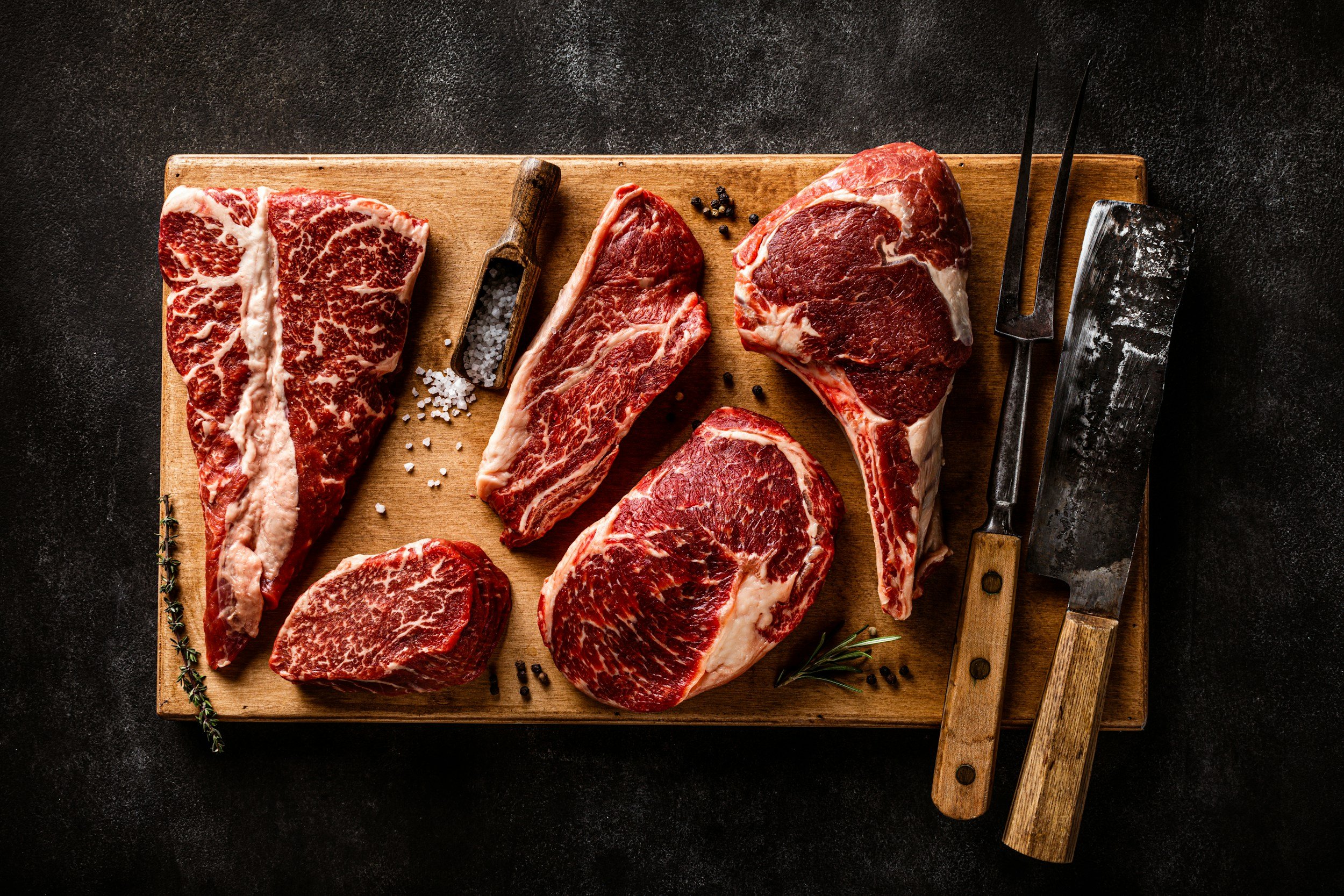 Assorted premium cuts of beef on a wooden cutting board, including ribeye, sirloin, and filet mignon, accompanied by a wooden scoop of salt and herbs. Ideal for gourmet cooking and grilling.